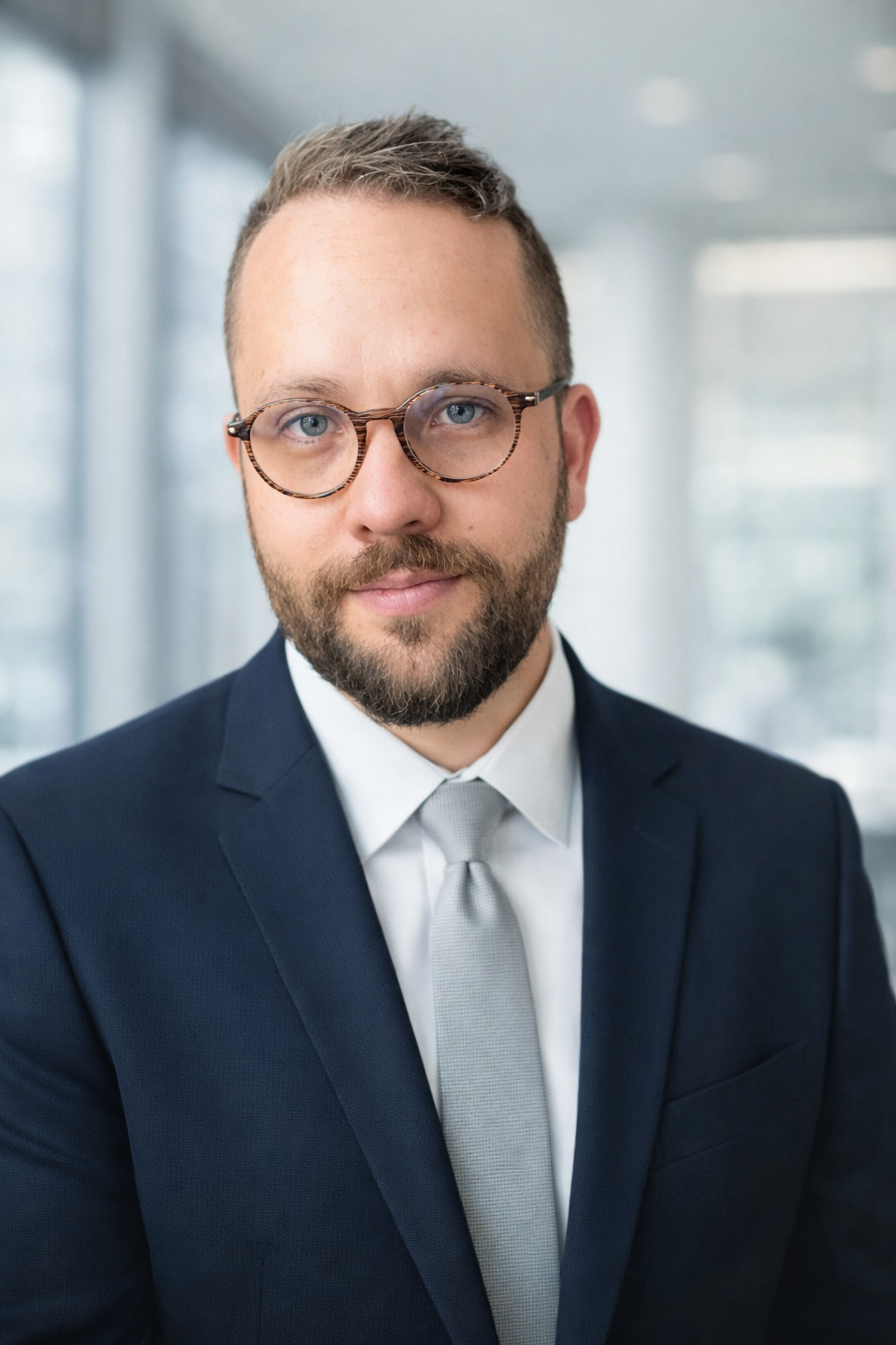 Adrian Carver, a man with short brown hair, a brown beard, and glasses wearing a navy suit with a grey tie